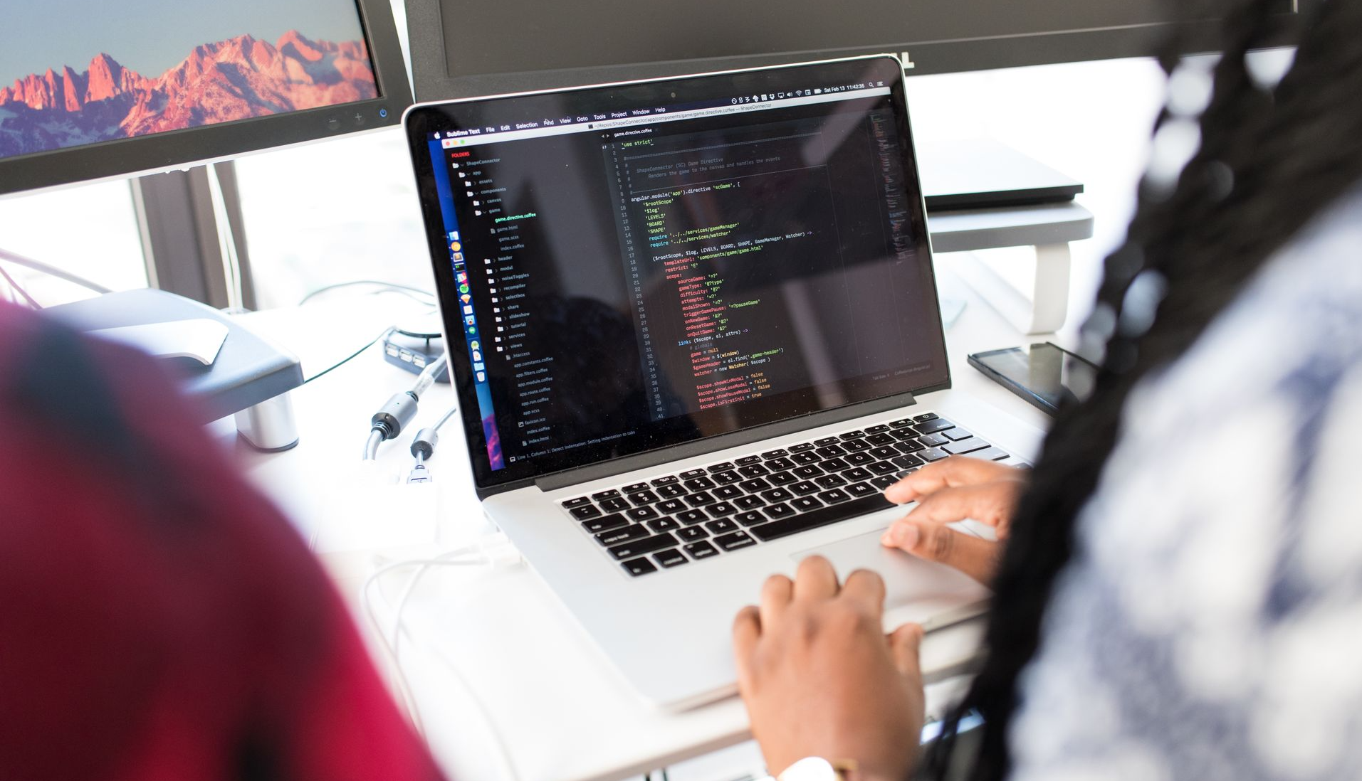 A woman coding on a laptop in a modern office environment with multiple monitors.