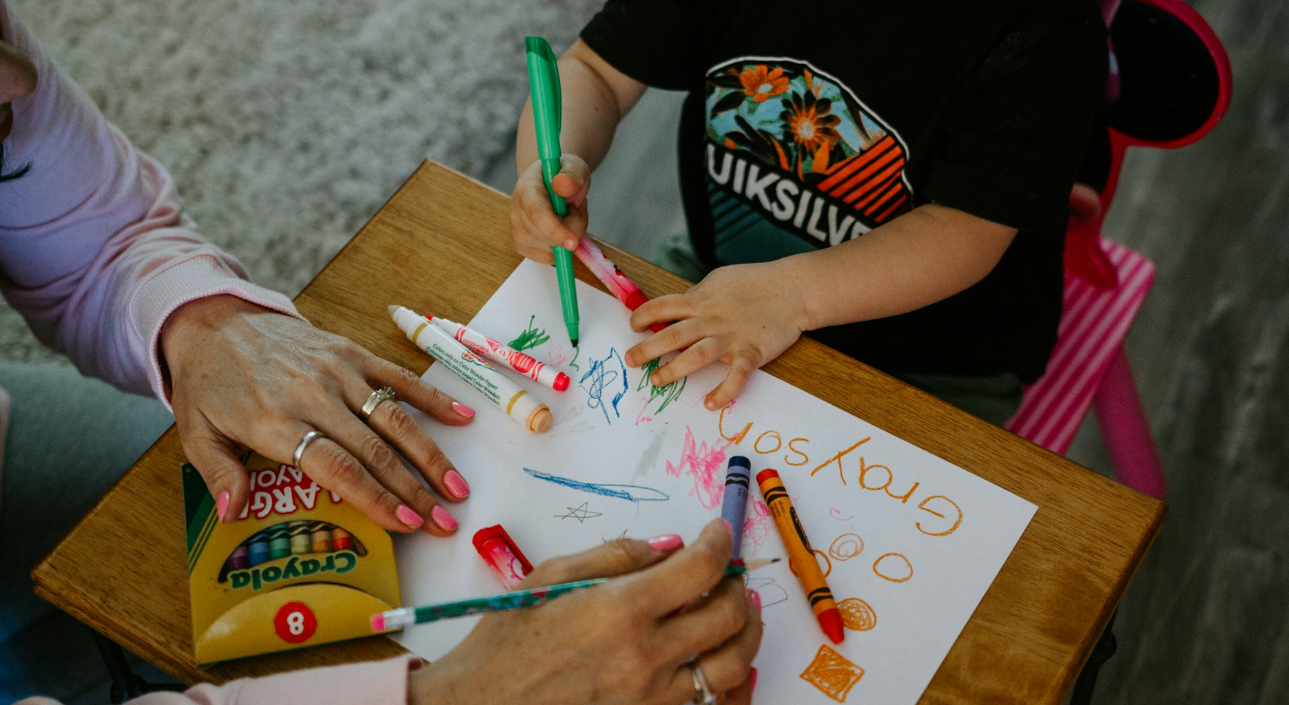 A woman helps a child color on a sheet of paper.