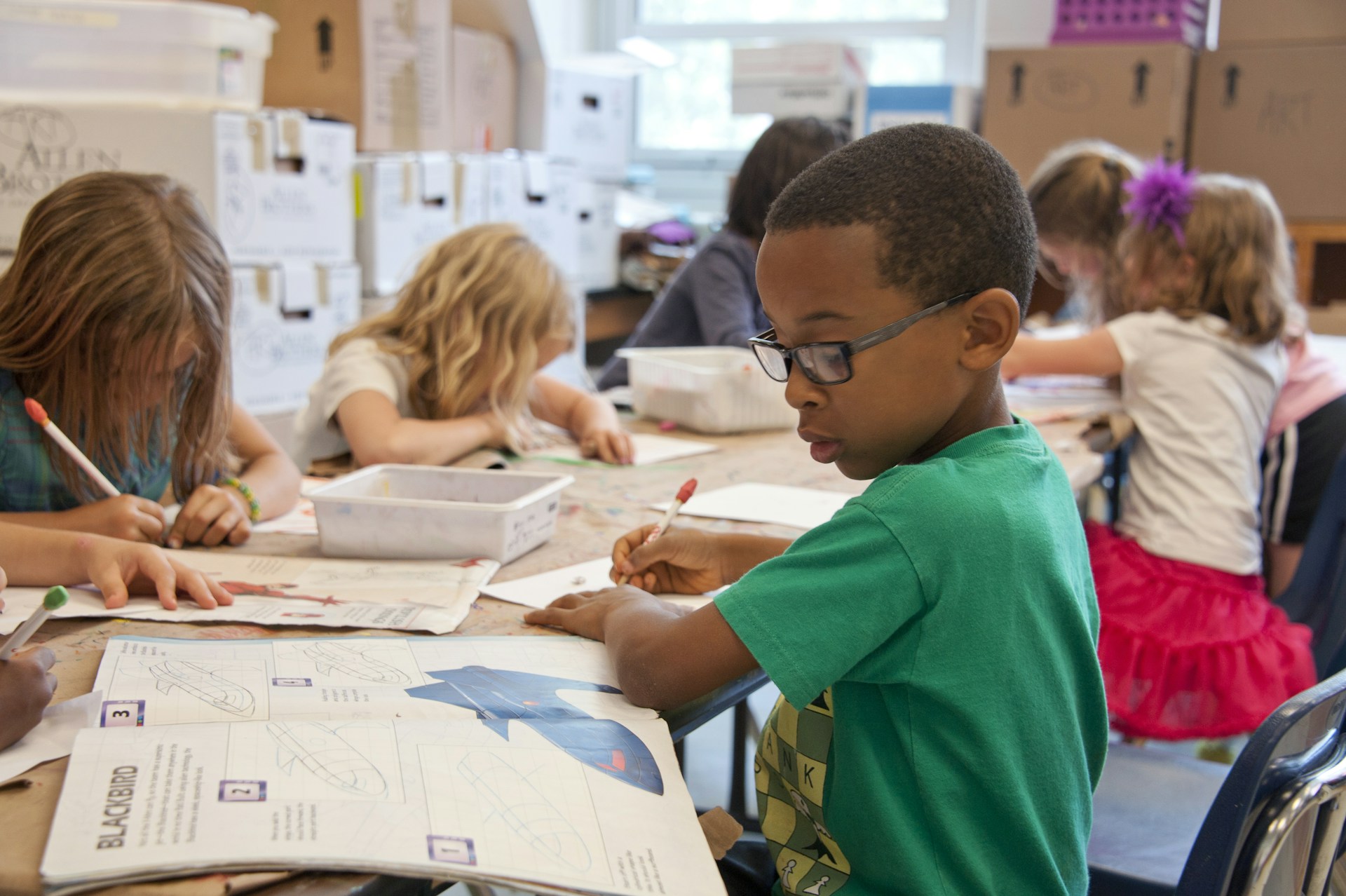 Various children at a table do their homework out of a workbook.